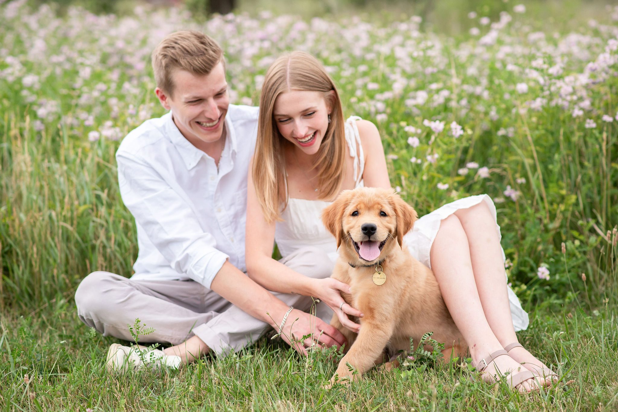 Couple sitting in grass smiling down at their 12 week old golden retriever puppy.