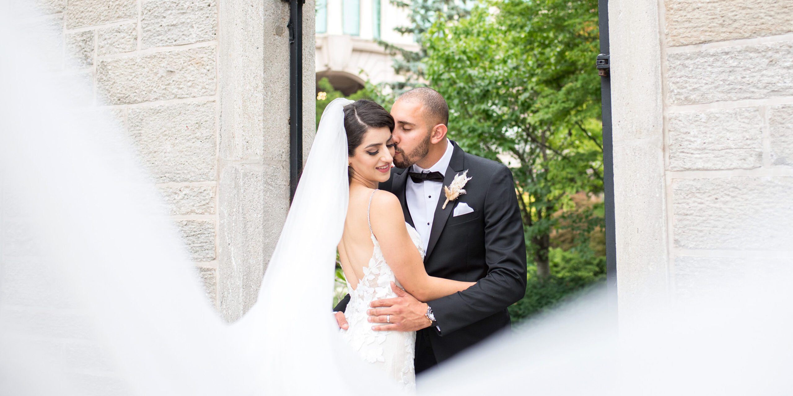 Groom kissing brides cheek as her veil flows in the foreground in Niagara Falls