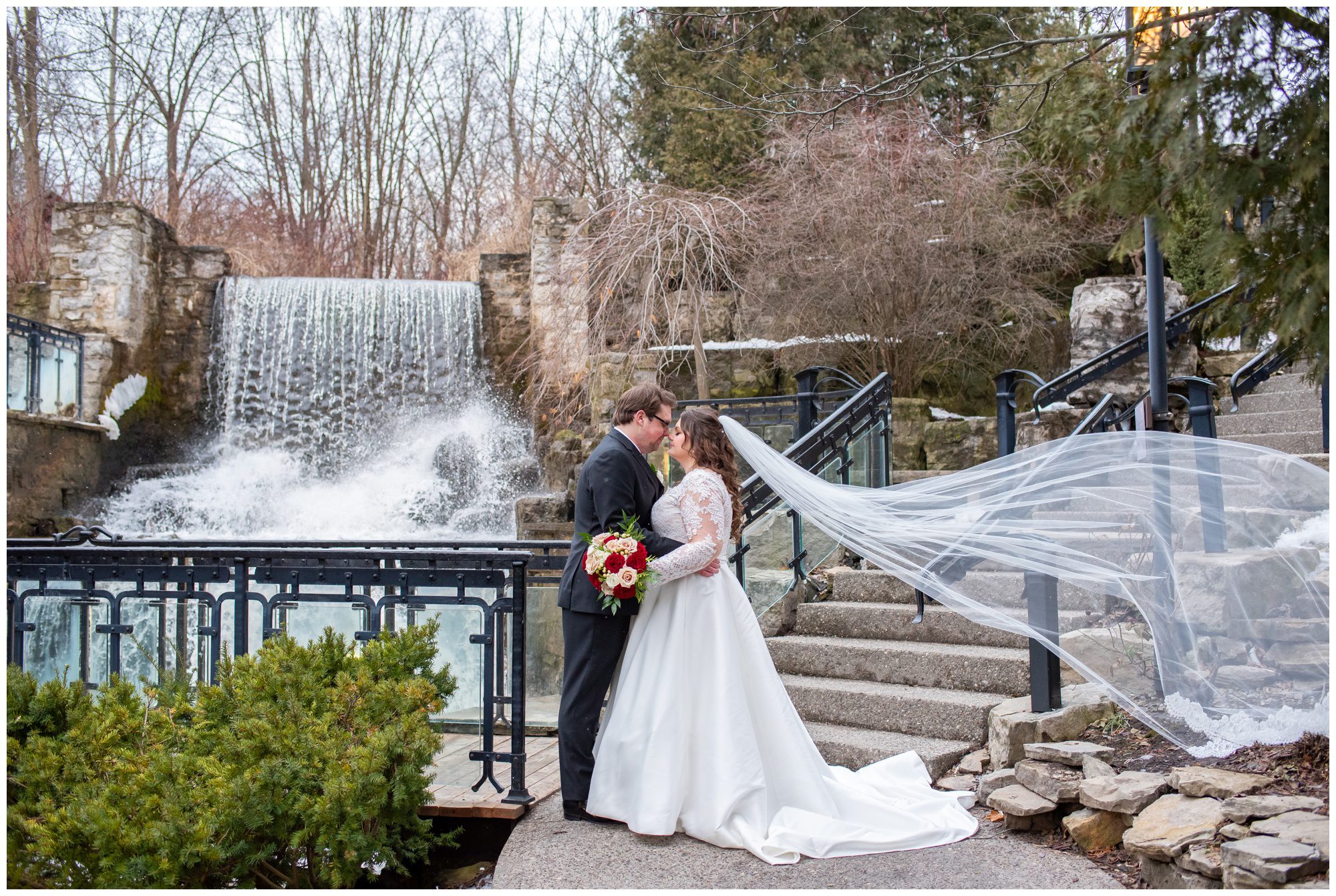 Bride and groom facing each other with veil flying in front of waterfall at Ancaster Mill.